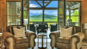 A cozy seating area with two cushioned chairs and a small table, a large window offering a scenic view of green hills, and a ceiling fan above. - 4 BR w Views, Private trails, hot tub, 3 king beds, EV Charger (Mineral Bluff)