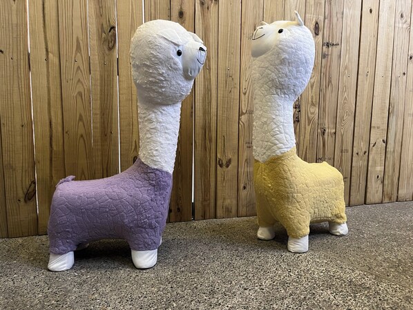 Two plush toys, one purple and one yellow, standing on a speckled floor against a wooden background.