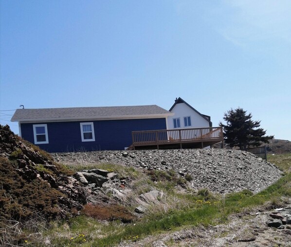 A house with a blue exterior, a wooden deck, and a rocky landscape. - Oceanview Cottage (Twillingate)