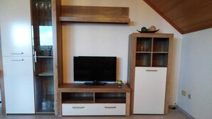 Una habitación con un mueble de entretenimiento de madera, un televisor de pantalla plana, un refrigerador blanco y un aparador de madera con puertas de vidrio.