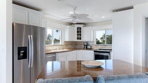 A modern kitchen with white cabinetry, a stainless steel refrigerator, a large island with a bowl, and a ceiling fan. - Private Beachfront House (Fort Pierce)