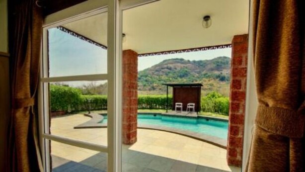 A view from a room with a large window overlooking a swimming pool, a brick wall, and a mountain in the background. - Royal Villa with Private pool (Lonavala)