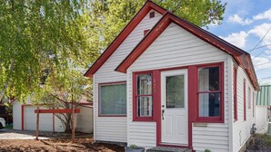 Une petite maison de plain-pied avec une porte et des fenêtres rouges, entourée d’un jardin agrémenté de paillis et de plantes en pot.