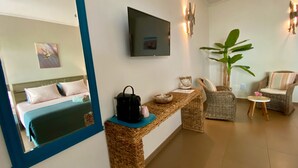 Une chambre d’hôtel comprenant un lit, un téléviseur fixé au mur, une console en osier, une chaise, une petite table avec un vase et une grande plante.