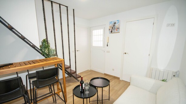 Un salon avec une table en bois, des chaises noires, un canapé blanc et une petite table ronde sur laquelle se trouvent une bouteille et un verre.