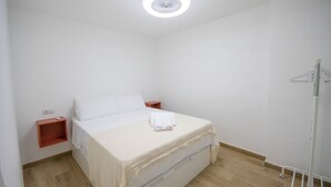 Un lit simple avec un matelas et une couverture blancs, une table de chevet, une petite étagère et un luminaire au plafond.