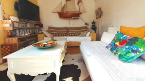 A cozy living room with a white sofa, a colorful cushion, a flat-screen TV, a bookshelf, and a sailboat decoration. - Very pleasant family home in Saint-Enogat Dinard (Dinard)
