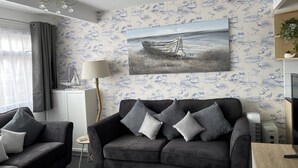 A living room with a grey sofa, a small side table, and a lamp. There is a painting of a boat on the wall. - M & T Chalets.  Refurbished Chalet sleeps 5, in Scratby , 5 mins to the beach. (great yarmouth)