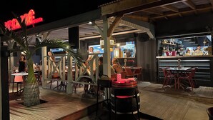 Un espace extérieur aménagé pour les repas, avec des tables et des chaises, un bar avec des étagères remplies de bouteilles, et un panneau lumineux au néon au-dessus de l’entrée.