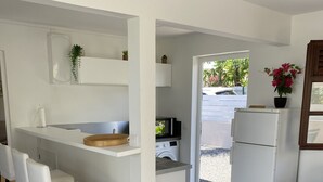 Une cuisine moderne avec des meubles blancs, un four à micro-ondes encastré et un lave-linge. On y trouve trois tabourets de bar blancs et une table en bois dans l’espace repas. Un canapé bleu est visible dans le salon.