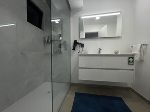 A modern bathroom with a glass shower enclosure, a white vanity with a mirror, and a blue bath mat. - Sea and Sun 4 You - Jardins da Imperatriz (Funchal)