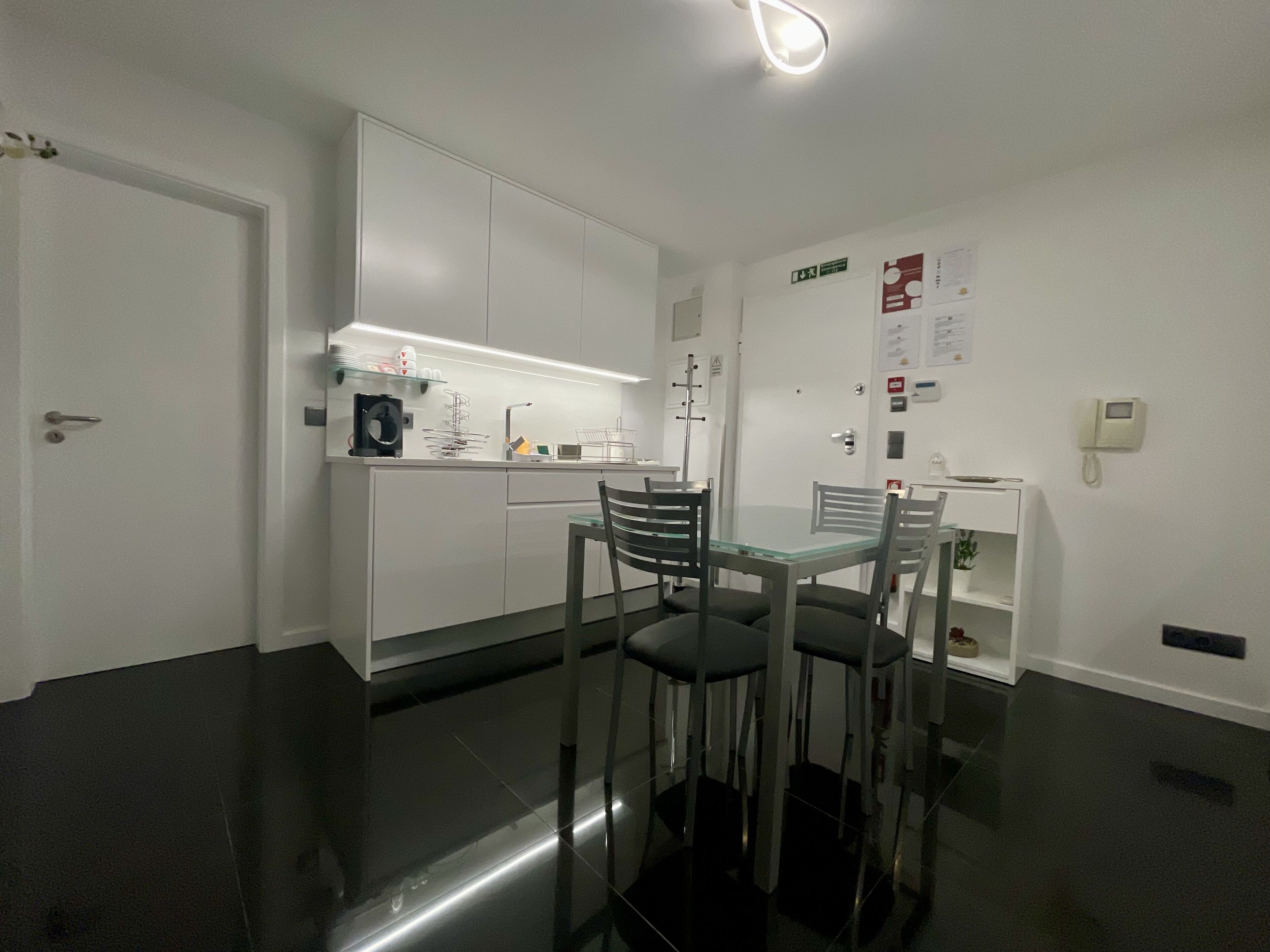 A modern kitchen with white cabinets, a glass dining table, and two chairs. There is a small island with a sink and a coffee machine.