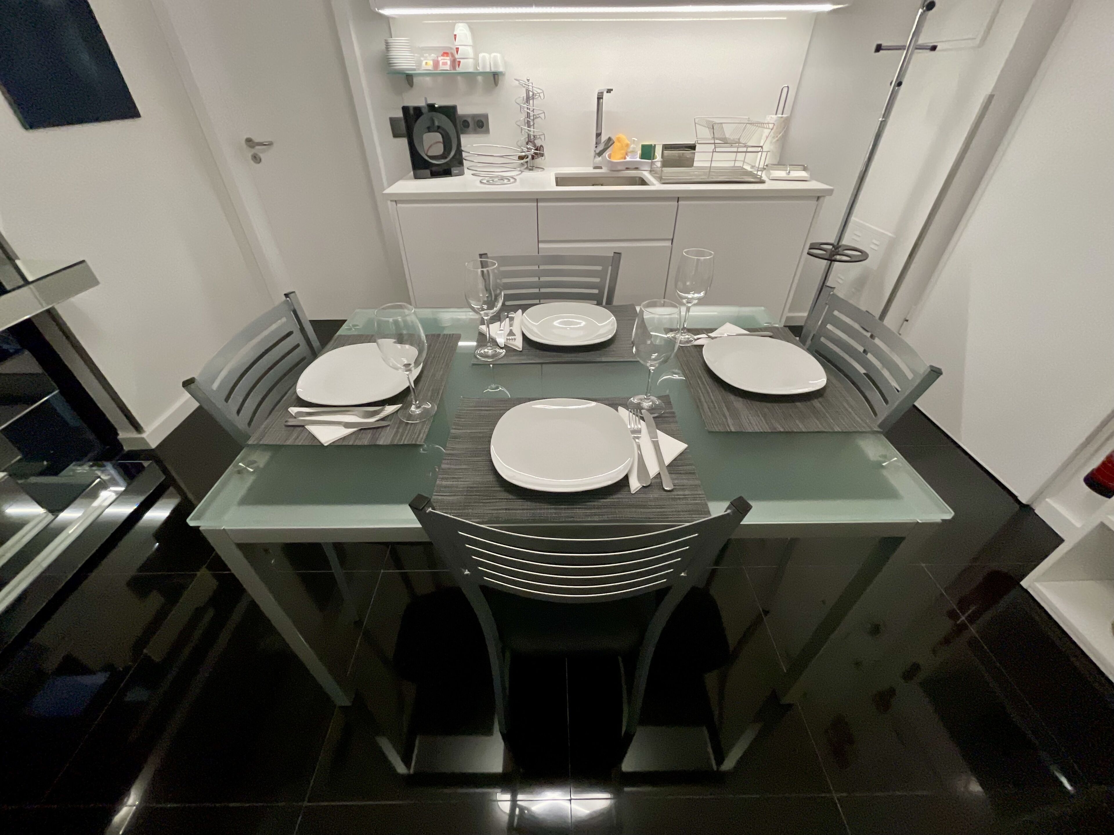 Et moderne spiseområde med et glasbord, metalstole og et køkken i baggrunden.