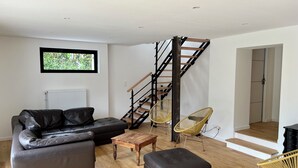 Uma sala de estar moderna com um sofá de couro, uma mesa de centro de madeira e um banquinho preto. Uma escada de madeira com corrimãos pretos leva a um segundo andar.