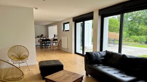 Uma sala de estar moderna com um sofá de couro, uma mesa de centro de madeira e uma cadeira de vime. Uma área de refeições é visível ao fundo, com uma mesa e cadeiras.
