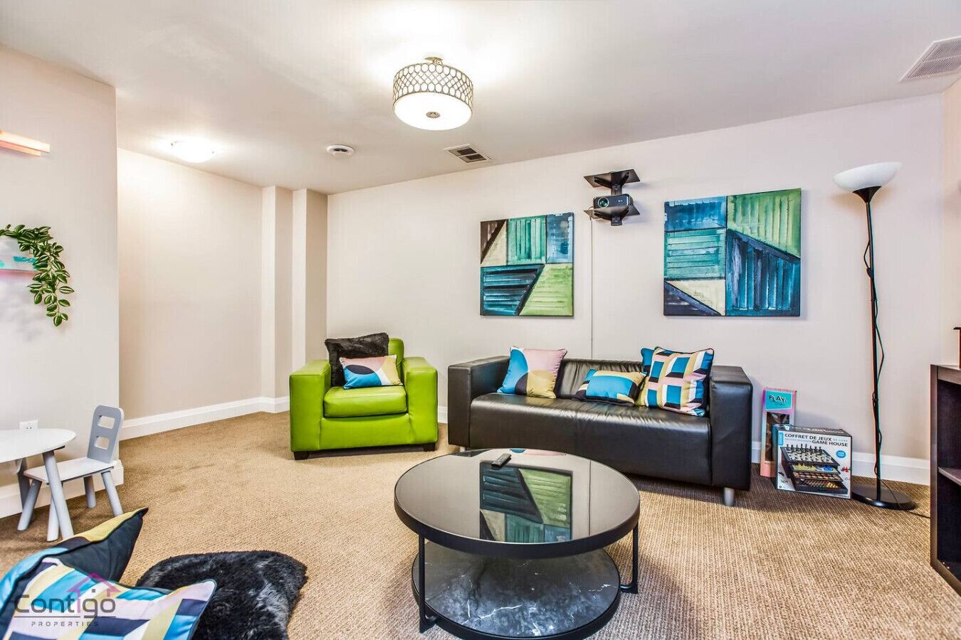 A modern living room with a green armchair, a black sofa, and a glass coffee table. There are abstract paintings on the wall and a lamp on the right.