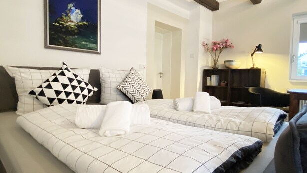 Habitación de hotel con dos camas, cada una con colcha y almohadas a cuadros, un cuadro en la pared, un escritorio con lámpara y ventana con vistas.