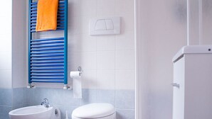 Uma casa de banho com duas pias, uma sanita e um porta-toalhas com uma toalha laranja, e um interruptor de luz na parede.