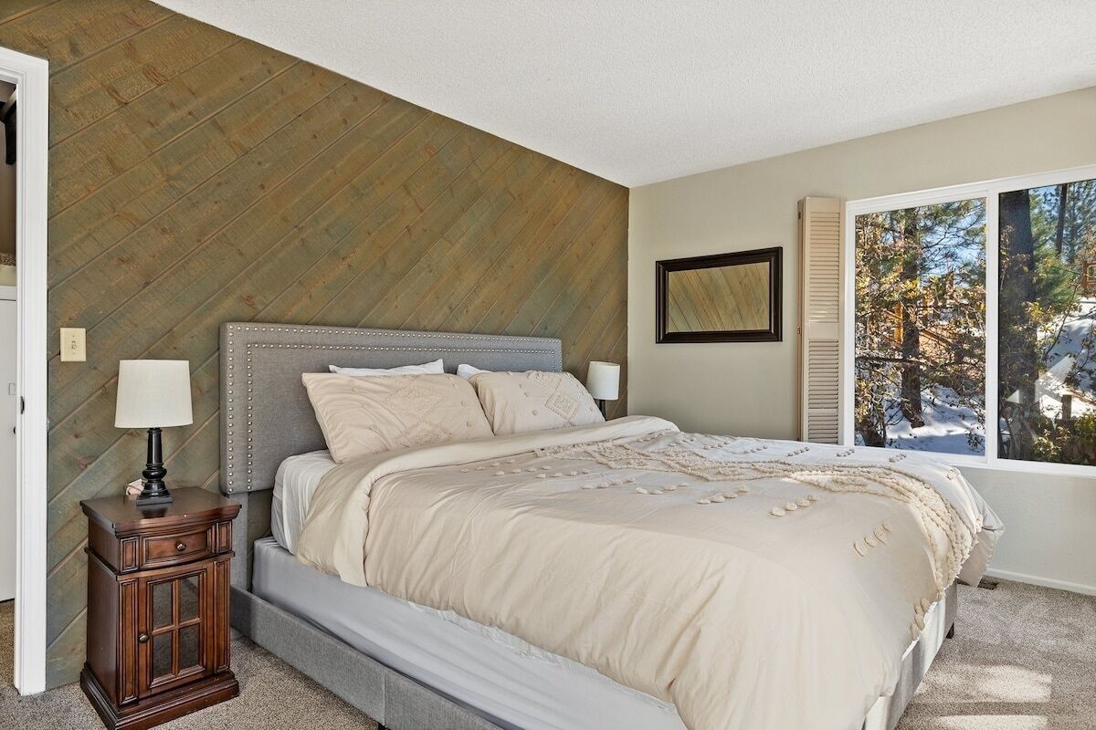 Un dormitorio con cama, una mesita de noche, una ventana con vistas a árboles y un panel de pared de madera.
