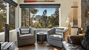 Un salon chaleureux avec deux fauteuils gris, un petit pouf rond et un canapé en cuir noir. Une grande fenêtre offre une vue sur les arbres et les montagnes.