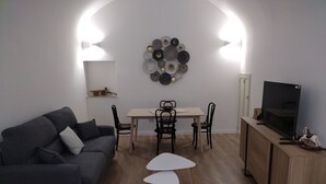 Een moderne woonkamer met een grijze bank, een houten eettafel met vier stoelen, een tv op een houten kast, en een wanddecoratie van borden.