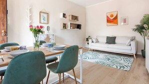 Una sala moderna con un sofá blanco, una mesa de comedor con sillas y una alfombra verde. Hay estanterías fijas a la pared y una planta en maceta.