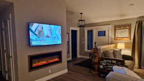 A living room with a flat-screen TV mounted on the wall, a fireplace below it, a couch, a coffee table, and a lamp. - Rainforest Getaway - Hot tub, bar, game room, creek in Forks...relax in style! (Forks)