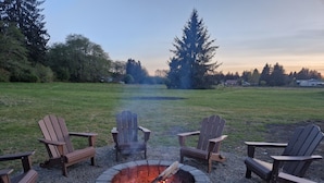 A fire pit with four Adirondack chairs around it, set on a gravel area with a grassy field in the background. - Rainforest Getaway - Hot tub, bar, game room, creek in Forks...relax in style! (Forks)