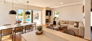 A modern living room with a beige sofa, a dining area with a table and chairs, a fireplace, and a pool visible through the glass doors.