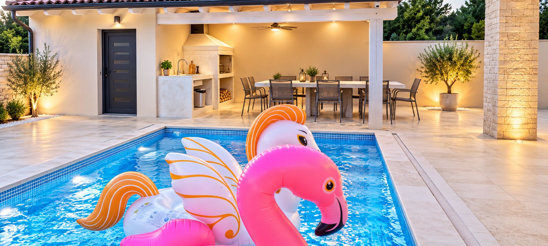Ein Swimmingpool mit aufblasbaren Flamingo-Schwimmfiguren, ein Essbereich mit Stühlen und Tischen sowie eine überdachte Terrasse mit Deckenventilator.