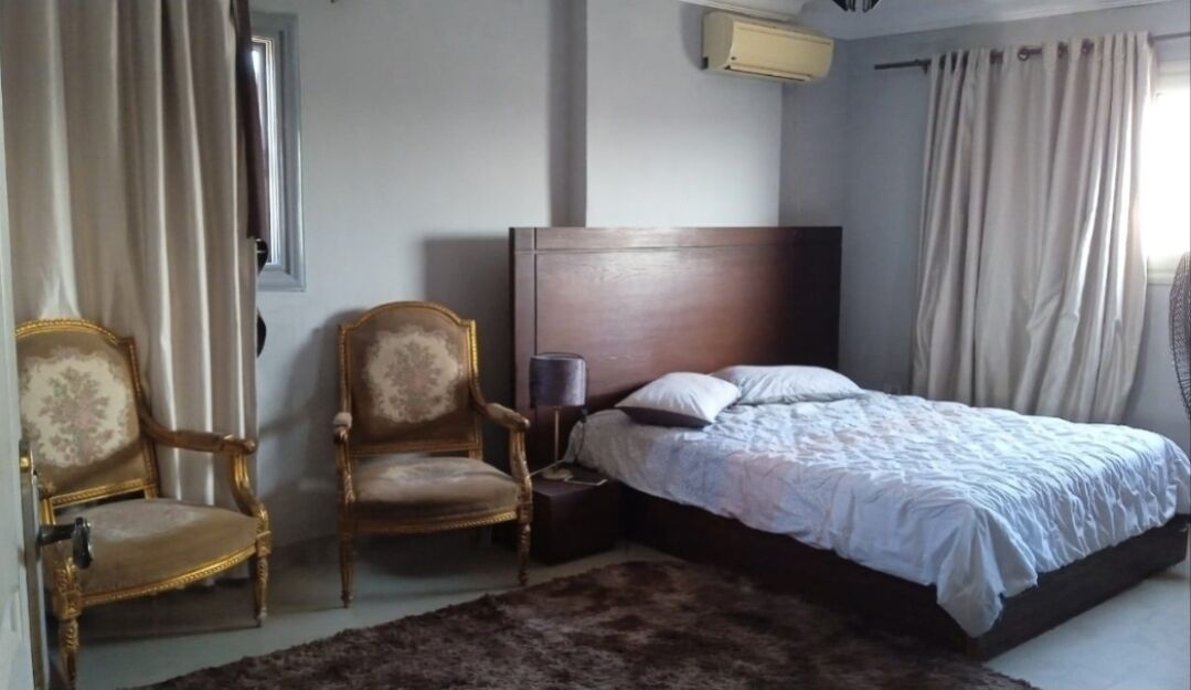 A bedroom with a bed, two armchairs, a nightstand, and a window with curtains.