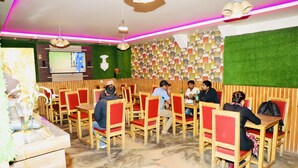Un restaurante con mesas de madera y sillas rojas con acolchado, un televisor de pantalla plana montado en la pared, y una pared verde con un diseño geométrico.