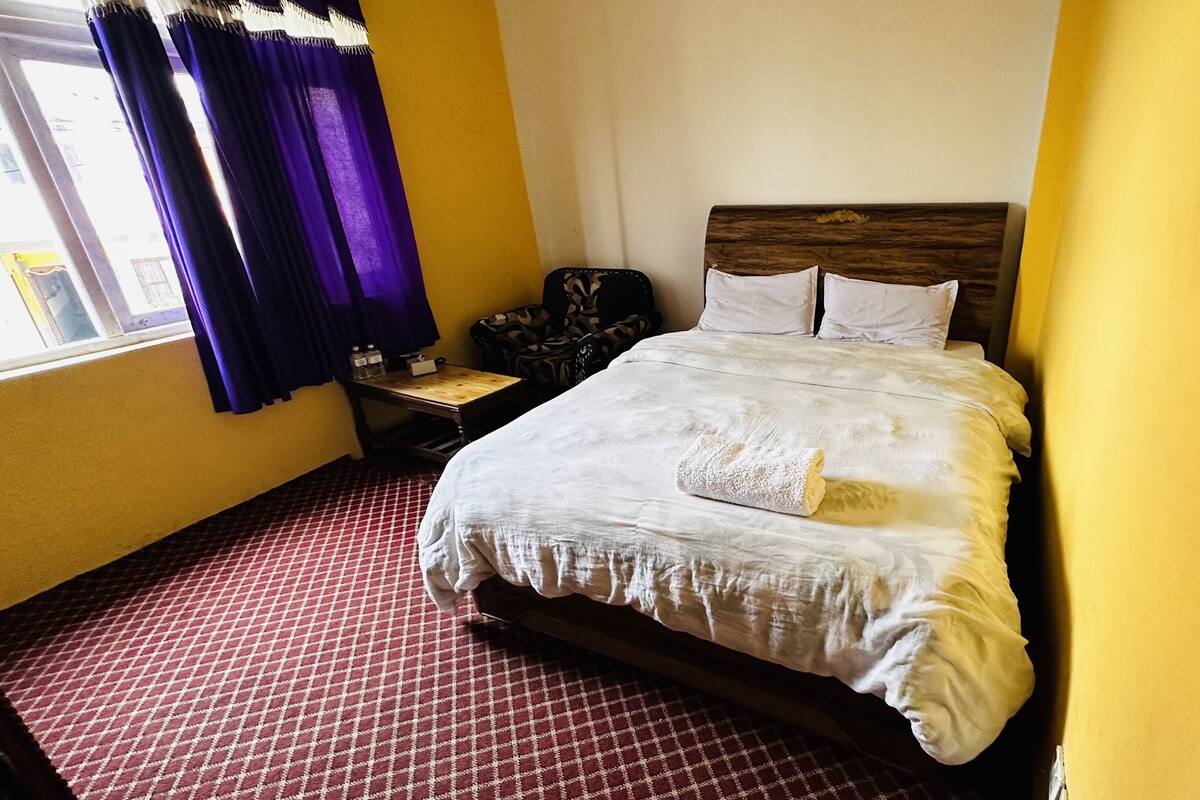 Una habitación de hotel con cama, mesita de noche, silla y ventana con cortinas.