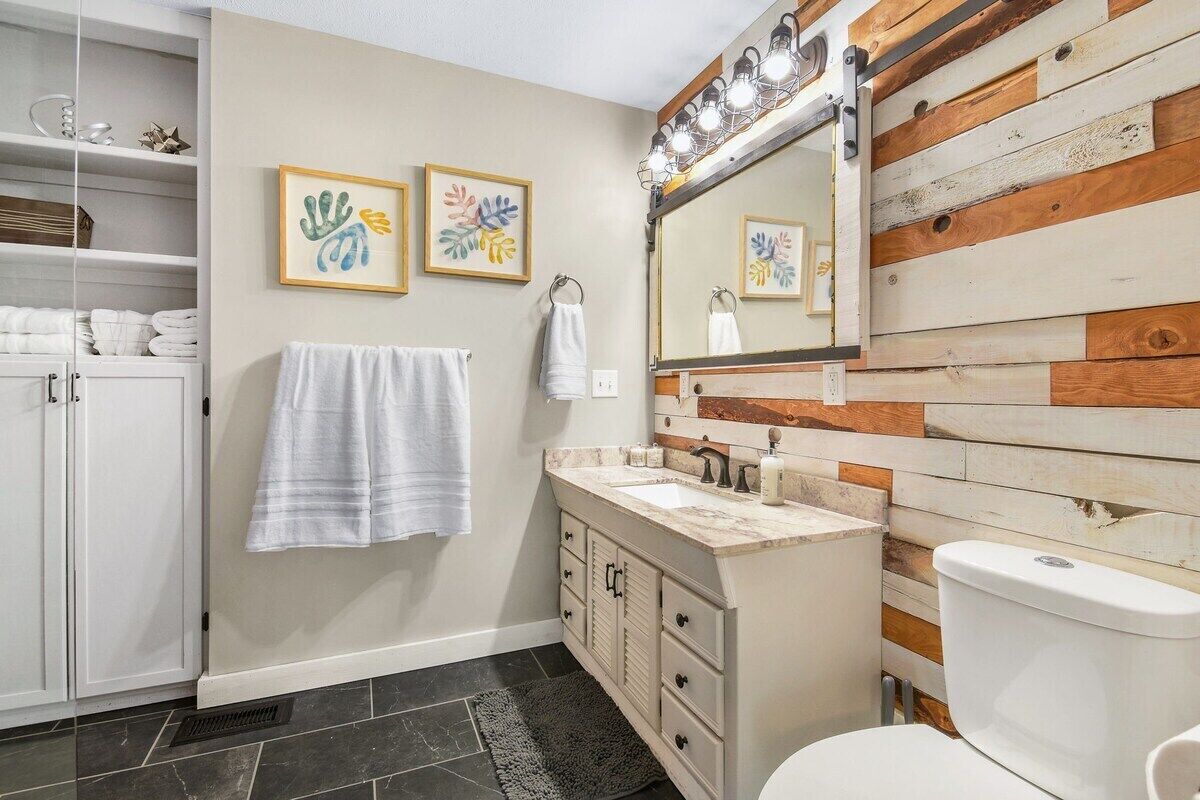 Une salle de bain avec un mur d’accent en bois, un meuble-lavabo blanc, un évier, des toilettes et deux œuvres d’art encadrées accrochées au mur.
