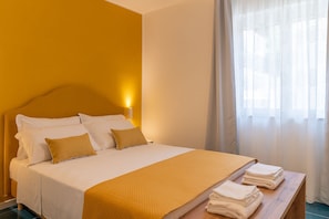 Ein Hotelzimmer mit Bett, Nachttisch mit Handtüchern, Kopfteil und einem Fenster mit Vorhängen.