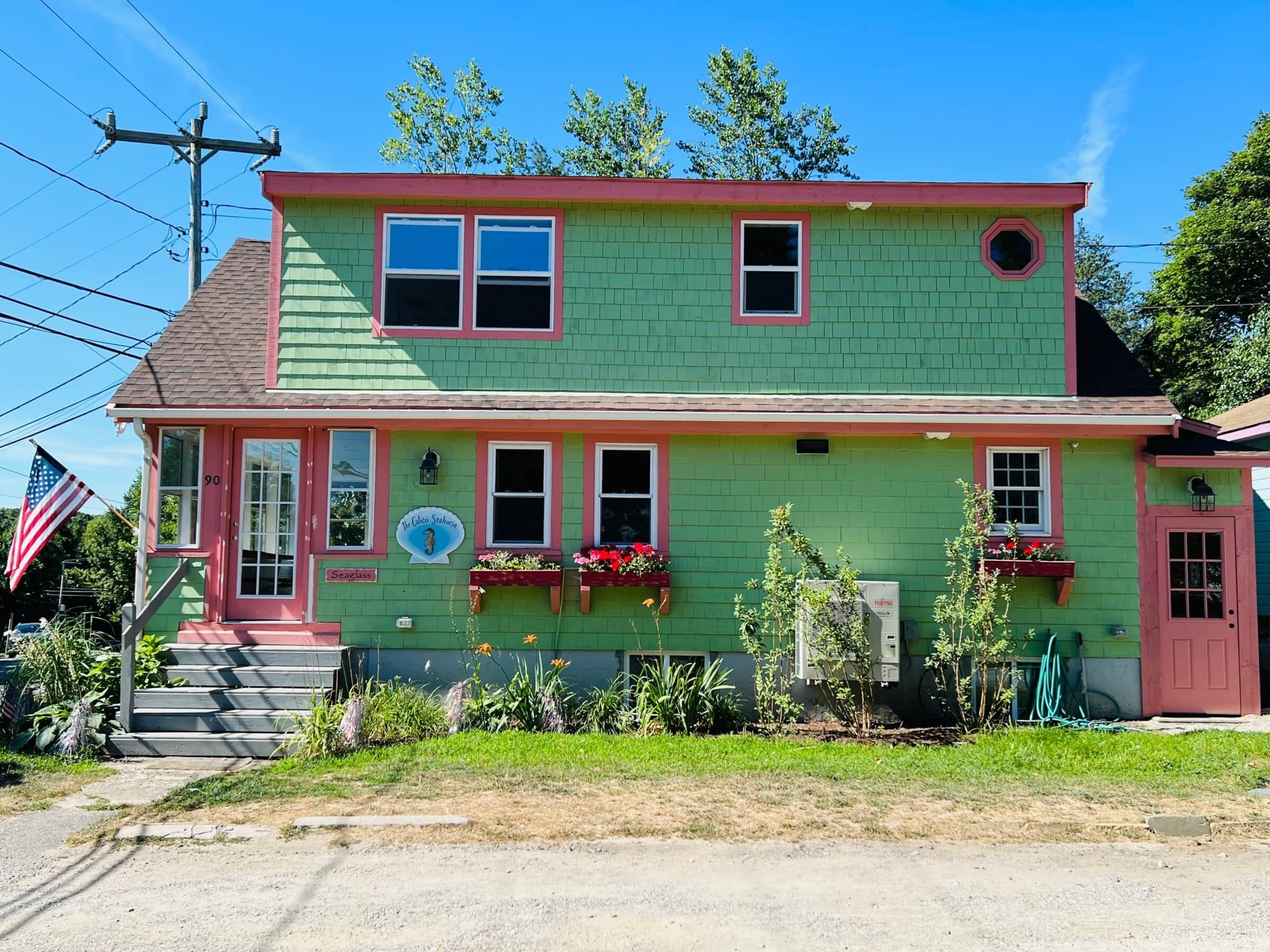 Uma casa de dois andares com fachada verde e detalhes vermelhos, apresentando um alpendre com degraus, uma caixa de correio e um pequeno jardim com flores.
