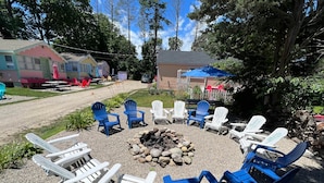 A fire pit surrounded by Adirondack chairs in a gravel area with a wooden deck and a building in the background. - Plymouth MA White Horse Beach Moana (Plymouth)