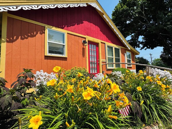 Una casa roja con detalles amarillos y una puerta roja, rodeada de flores y follaje amarillo.