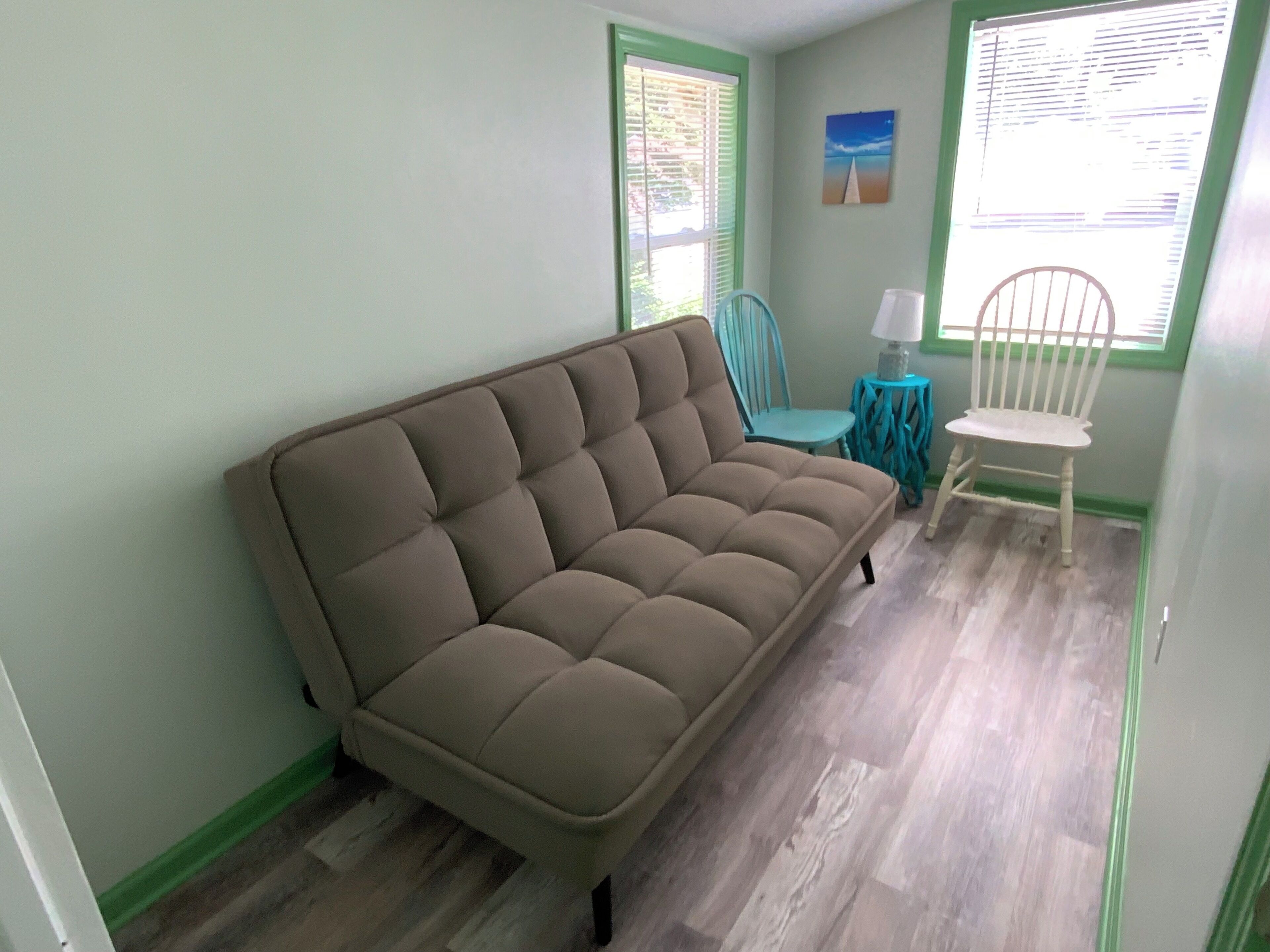 A room with a beige sofa, a small table with a lamp, a chair, and a window with blinds.