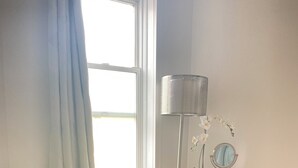 Ein Zimmer mit einem beigen Sofa, einem runden Holztischchen mit Spiegel und Zeitschrift, einer stehenden Lampe und einem Fenster mit Vorhängen.