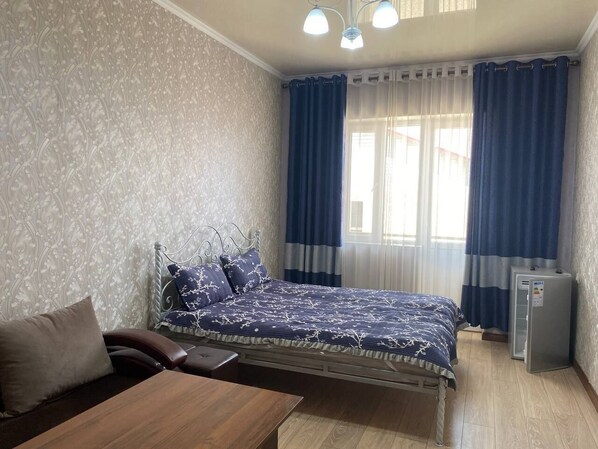 A bedroom with a bed, a small table, a chair, a desk, and a refrigerator.