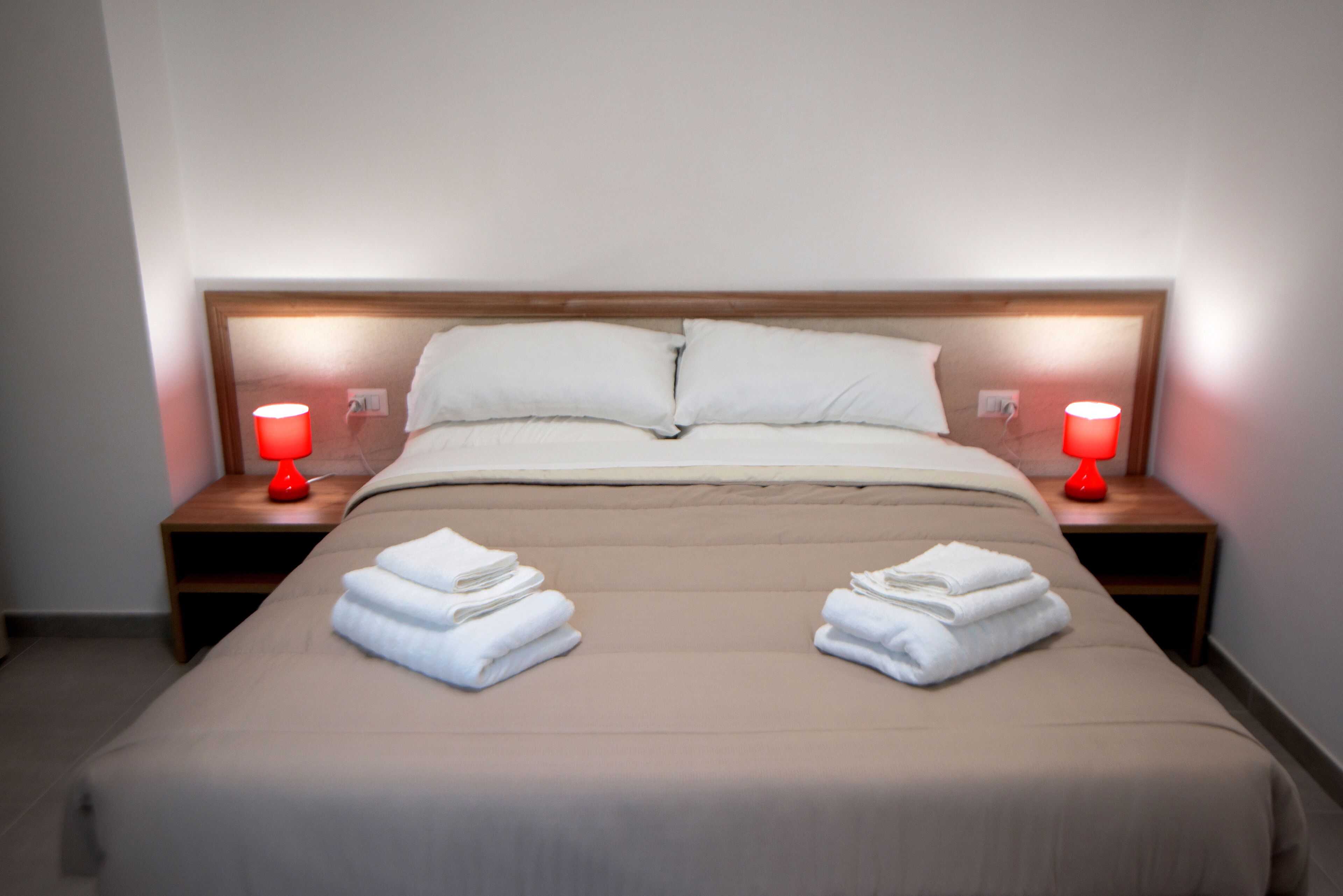 A neatly made bed with two pillows, two red bedside lamps, and folded towels on each side.