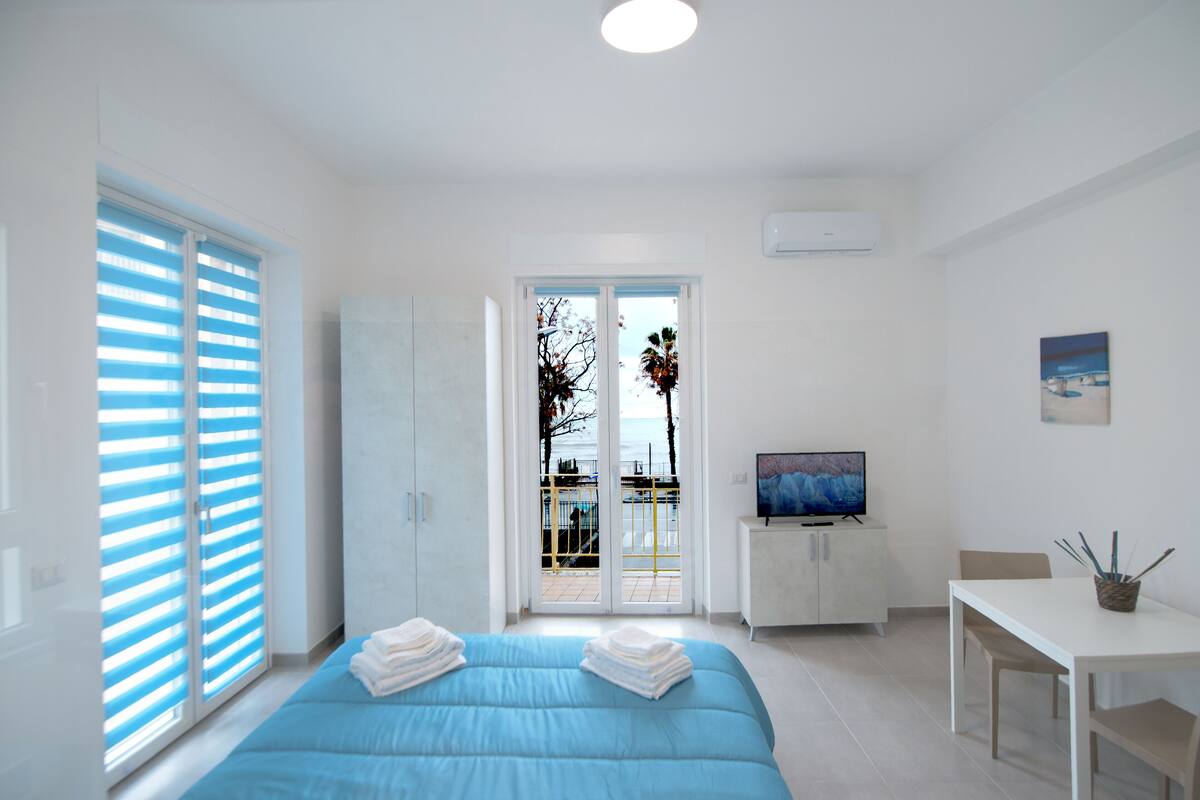 A modern bedroom with a bed, a TV, and a balcony with a view of the sea.