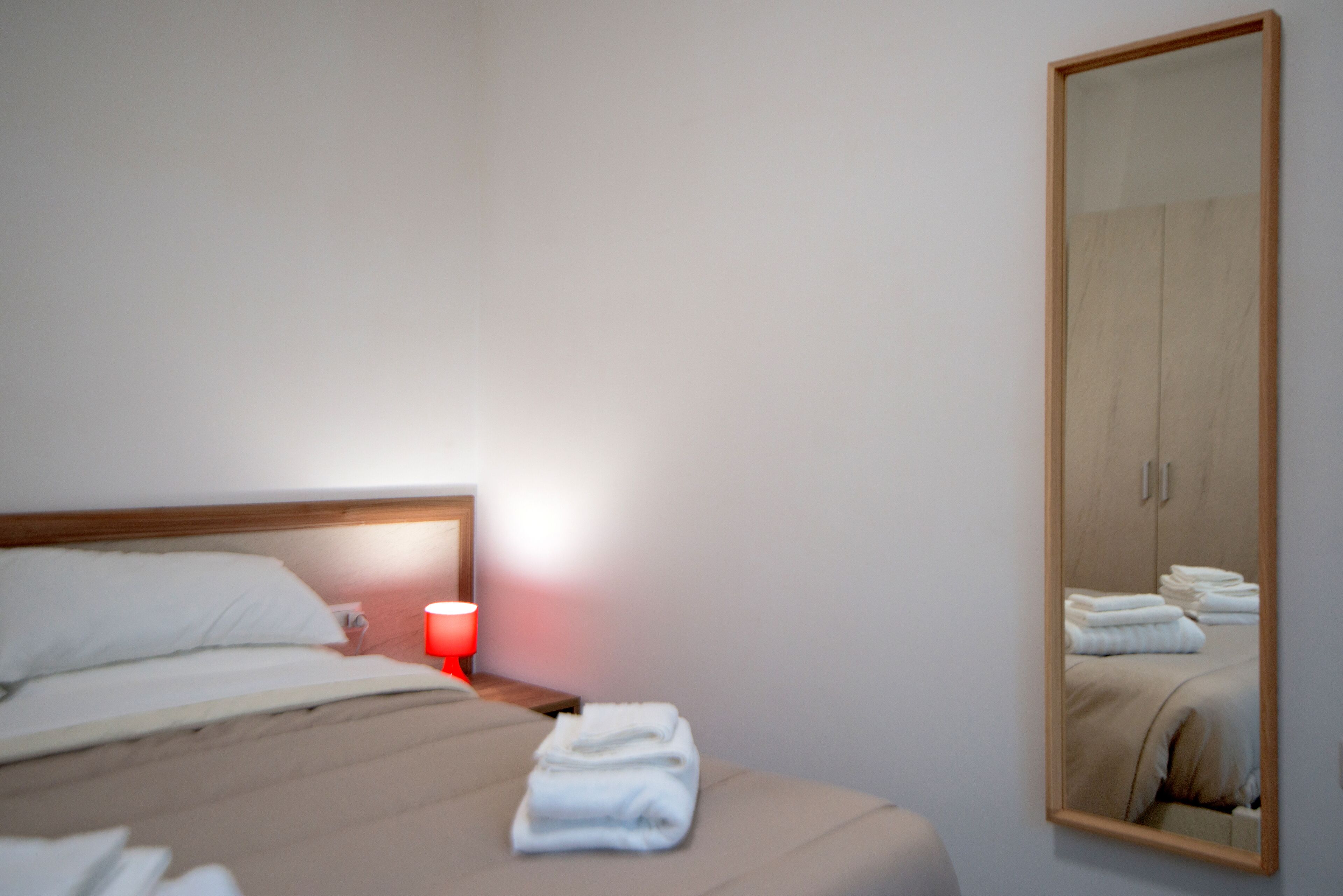 Een net opgemaakt bed met witte lakens, een rode bedlamp en een spiegel die de kamer weerspiegelt.