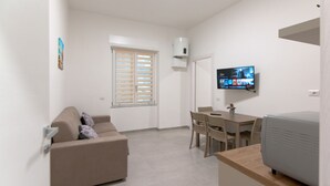 Una sala de estar moderna con un sofá color beige, una mesa de comedor con sillas, un televisor de pantalla plana en la pared y una pequeña área de cocina con un refrigerador.