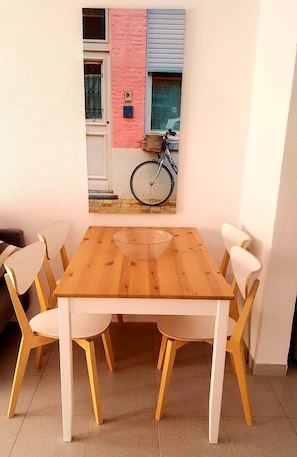 Un comedor con una mesa de madera y sillas blancas, un cuenco de vidrio sobre la mesa y una pared colorida con una ventana y una bicicleta.