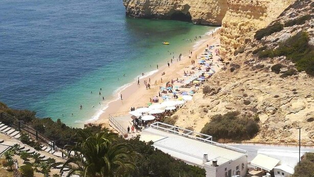 A coastal beach with a rocky cliff, a sandy shore, and a long row of beach umbrellas. - Chabella - Charm Beach Lofts & Apartments - Centeanes (Carvoeiro)
