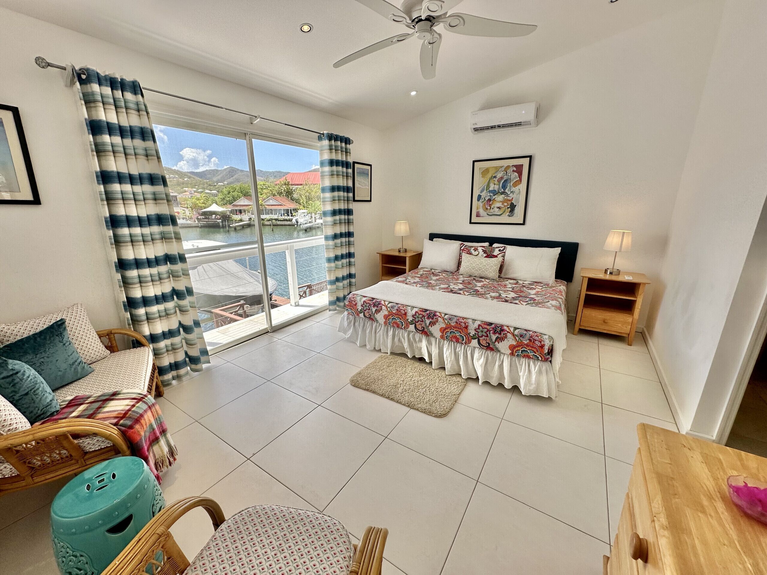 A bedroom with a bed, two bedside tables, a ceiling fan, an air conditioning unit, a window with curtains, and a view of a body of water and buildings.