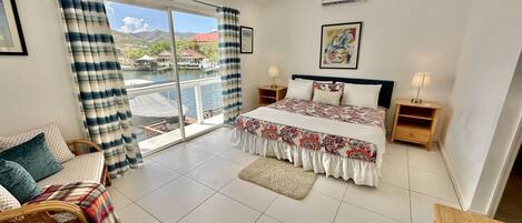 A bedroom with a bed, two bedside tables, a ceiling fan, an air conditioning unit, a window with curtains, and a view of a body of water and buildings.
