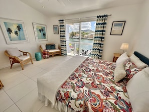 A bedroom with a large bed, a wicker chair, a sofa, a view of the water, and a balcony.